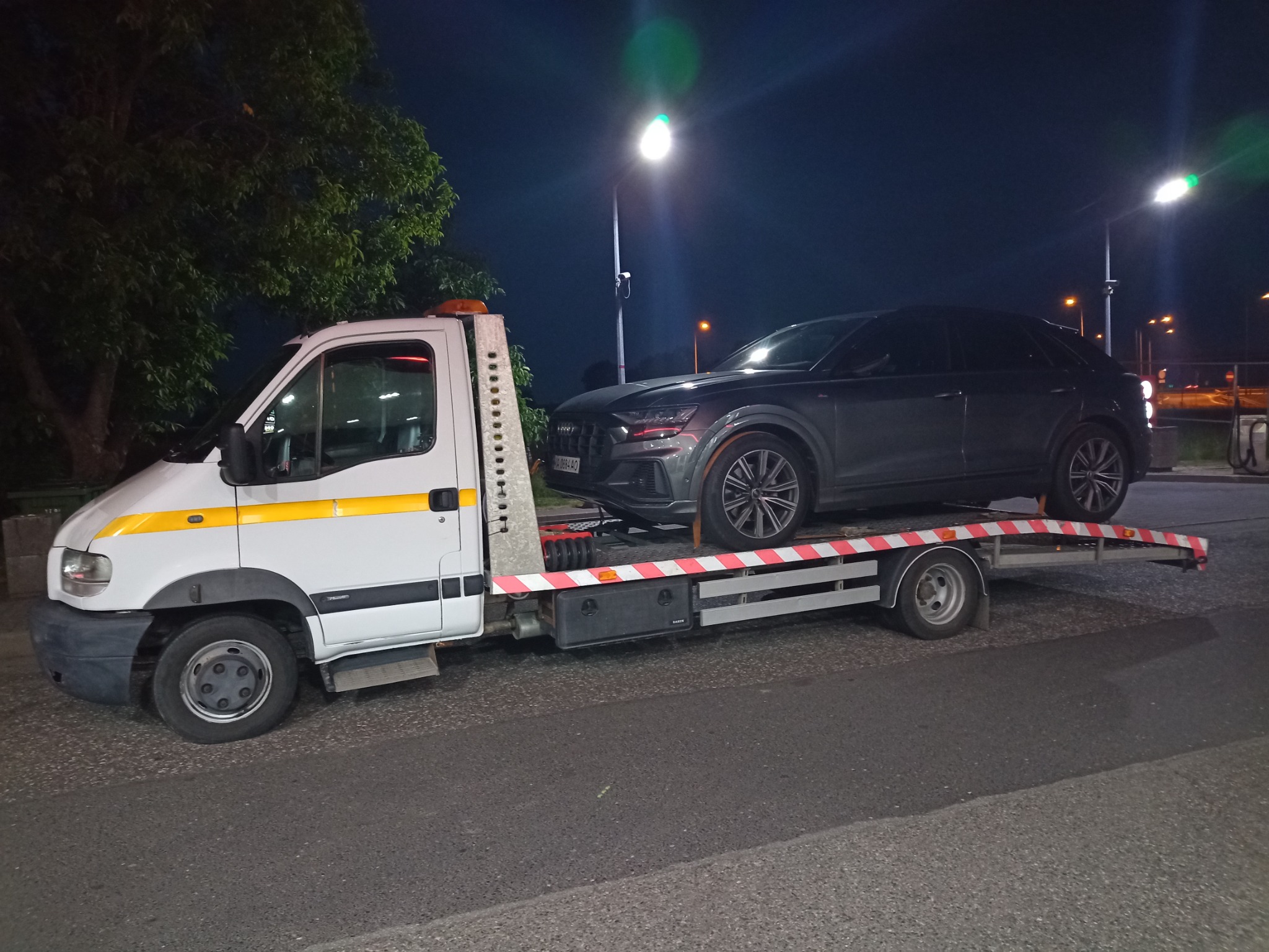 Ciemnoszary samochód osobowy Audi Q8 transportowany na platformie pojazdu pomocy drogowej w nocy, oświetlonego przez uliczne lampy.