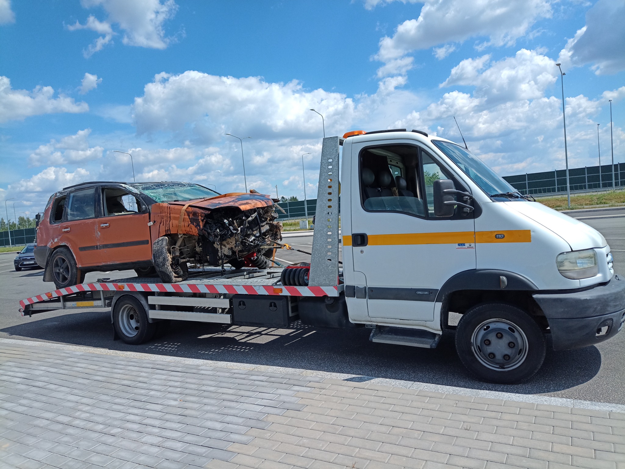 Pomarańczowy samochód osobowy po wypadku, z widocznymi uszkodzeniami karoserii i rozbitymi szybami, transportowany na platformie pojazdu pomocy drogowej w słoneczny dzień.