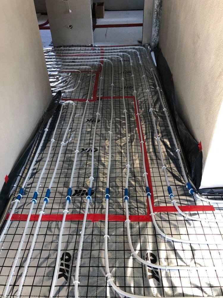 Rozłożona instalacja ogrzewania podłogowego z białych rur na folii aluminiowej z czerwoną siatką, widoczne niebieskie złączki i białe uchwyty mocujące rury do podłoża, surowy stan wykończenia wnętrza.