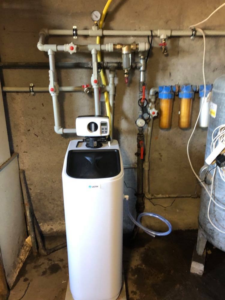 Biały zmiękczacz wody USTM podłączony do instalacji z rur PCV z czerwonymi zaworami, manometrem i filtrami narurowymi w pomieszczeniu technicznym.