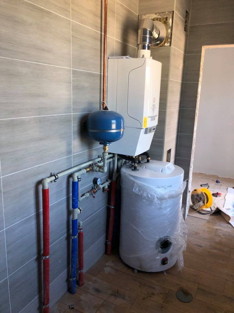 Nowo zainstalowany, biały, kondensacyjny kocioł gazowy z niebieskim naczyniem przeponowym i owiniętym folią ochronną, białym zasobnikiem ciepłej wody w pomieszczeniu z szarymi płytkami...