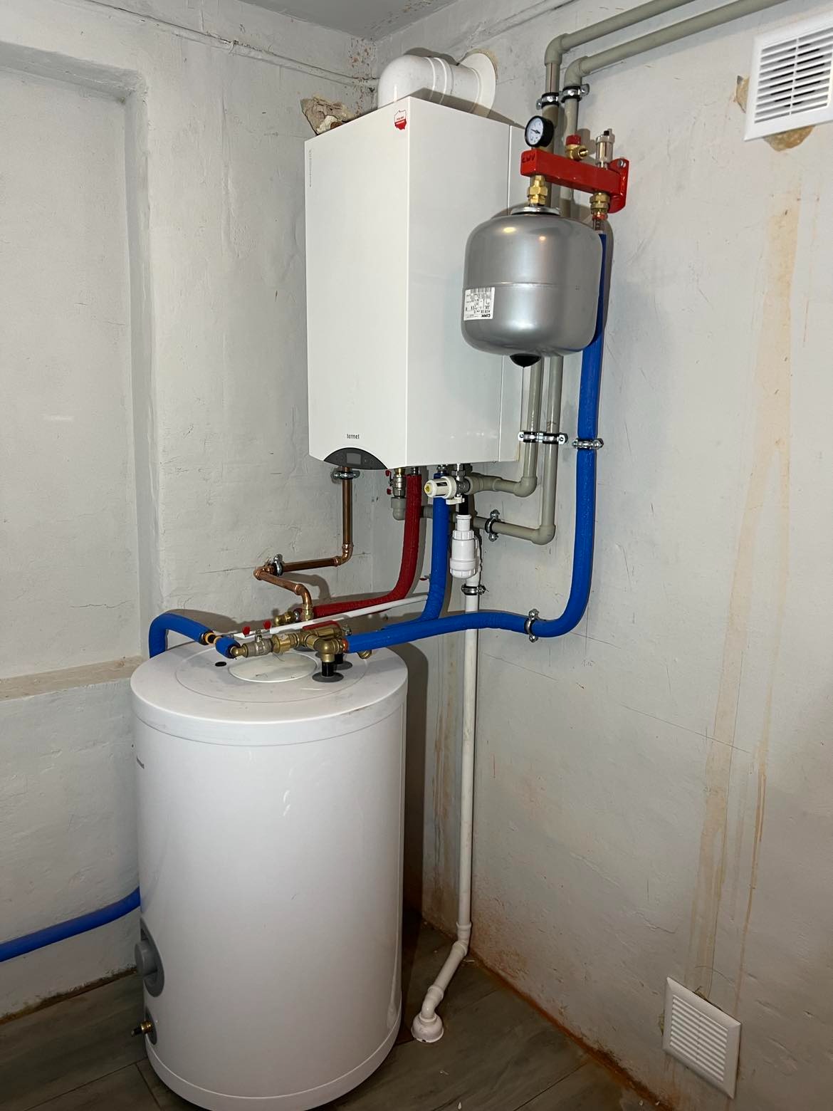 Biały piec gazowy Termet zamontowany na ścianie, połączony rurami miedzianymi i niebieskimi z białym zasobnikiem ciepłej wody, widoczne manometry i zawory, instalacja w surowym pomieszczeniu.