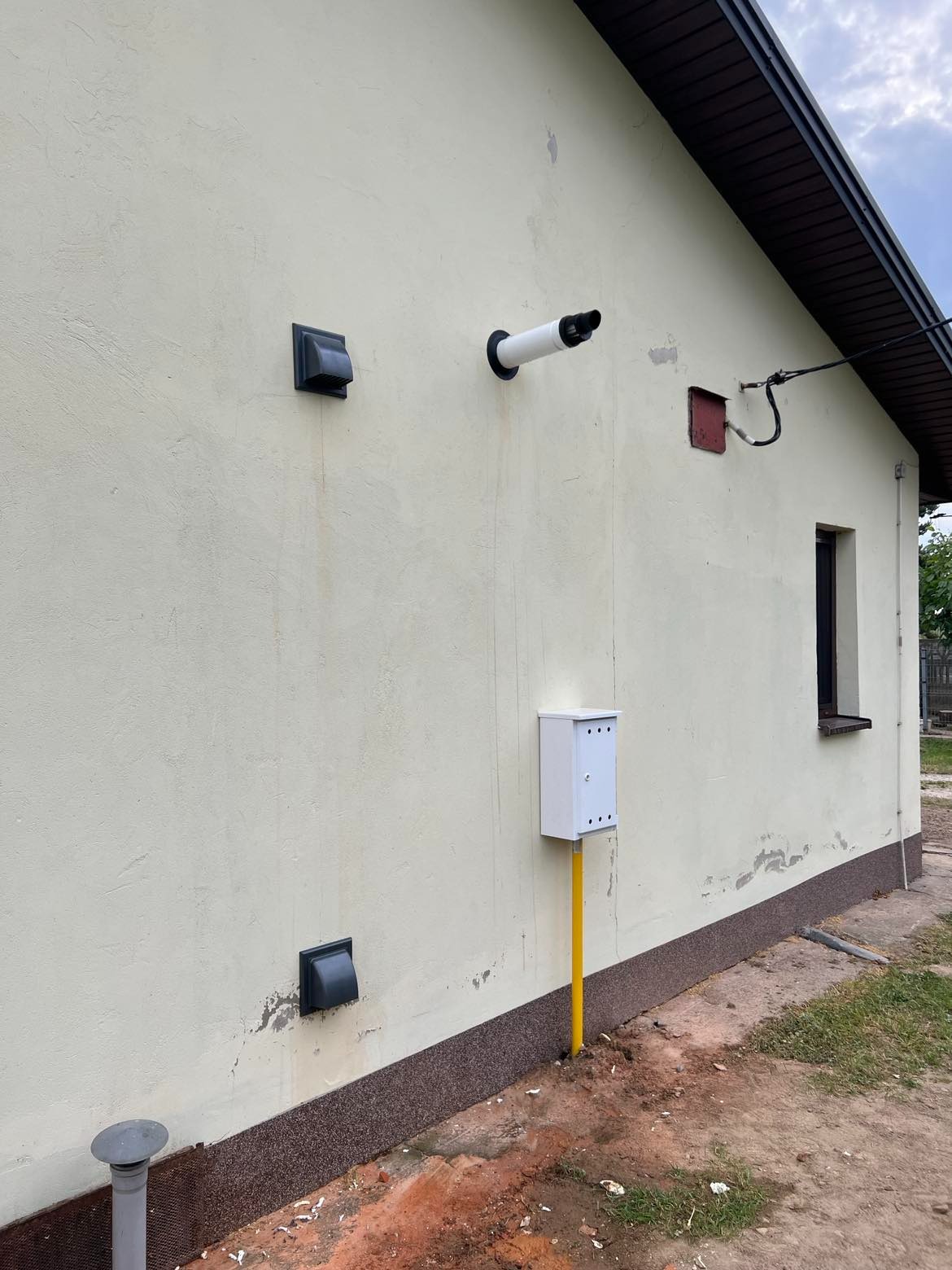 Elewacja budynku z widocznymi elementami instalacji: dwa czarne wywietrzniki, biała rura wentylacyjna, skrzynka gazowa na żółtej rurze i szara rura kanalizacyjna przy ziemi.