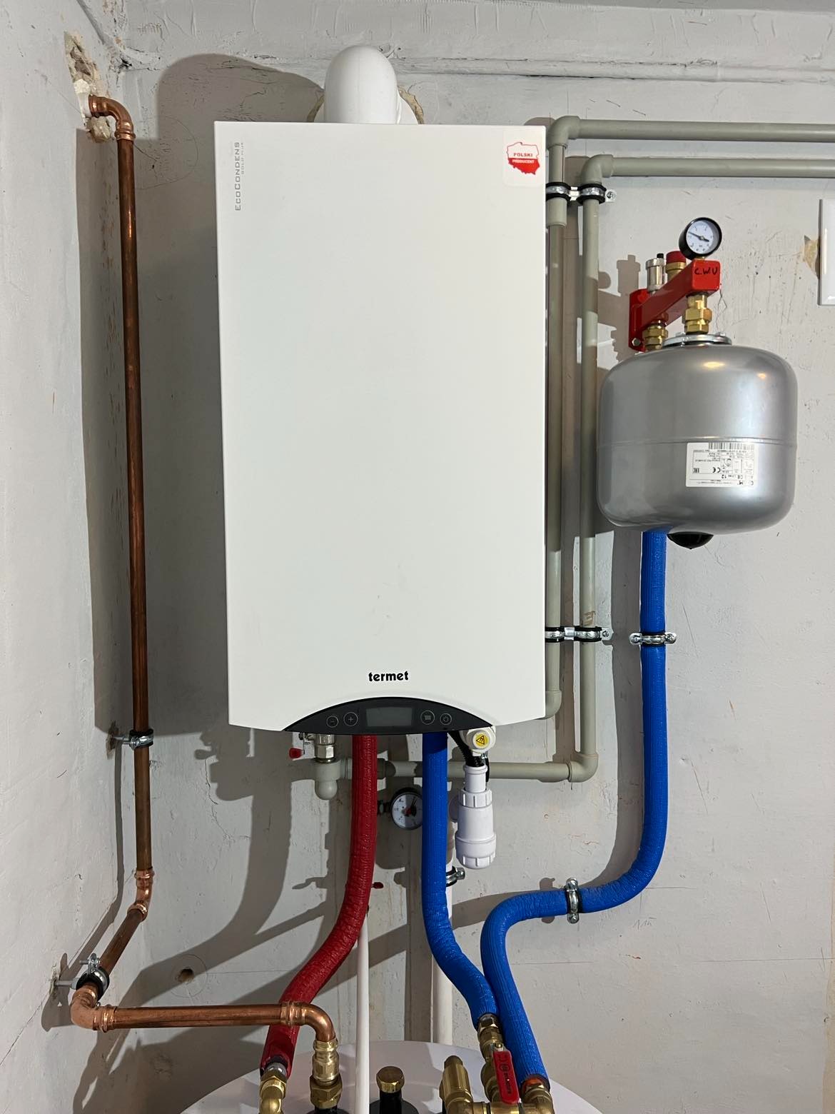 Biały, wiszący piec gazowy marki Termet z podłączonymi rurami miedzianymi i niebieskimi elastycznymi przewodami w surowym wnętrzu z widoczną instalacją.