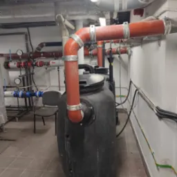 Czarny zbiornik z podłączonymi czerwonymi rurami w pomieszczeniu technicznym z widocznymi przewodami i instalacjami pod sufitem.