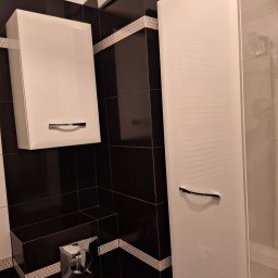 uslugi budowllane aksana hanchar - Ukośne ujęcie fragmentu łazienki z czarno-białymi płytkami, wiszącą białą szafką z chromowanym uchwytem, wysoką białą szafką przy prysznicu i chromowanym przyciskiem spłuczki.