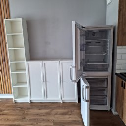 uslugi budowllane aksana hanchar - Częściowo opróżniona, biała lodówka z otwartymi drzwiami w mieszkaniu z drewnianą podłogą, białymi szafkami i półkami.