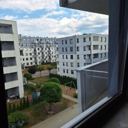 uslugi budowllane aksana hanchar - Widok z okna na osiedle bloków z placem zabaw i zielenią, częściowo przesłonięty ramą okienną.