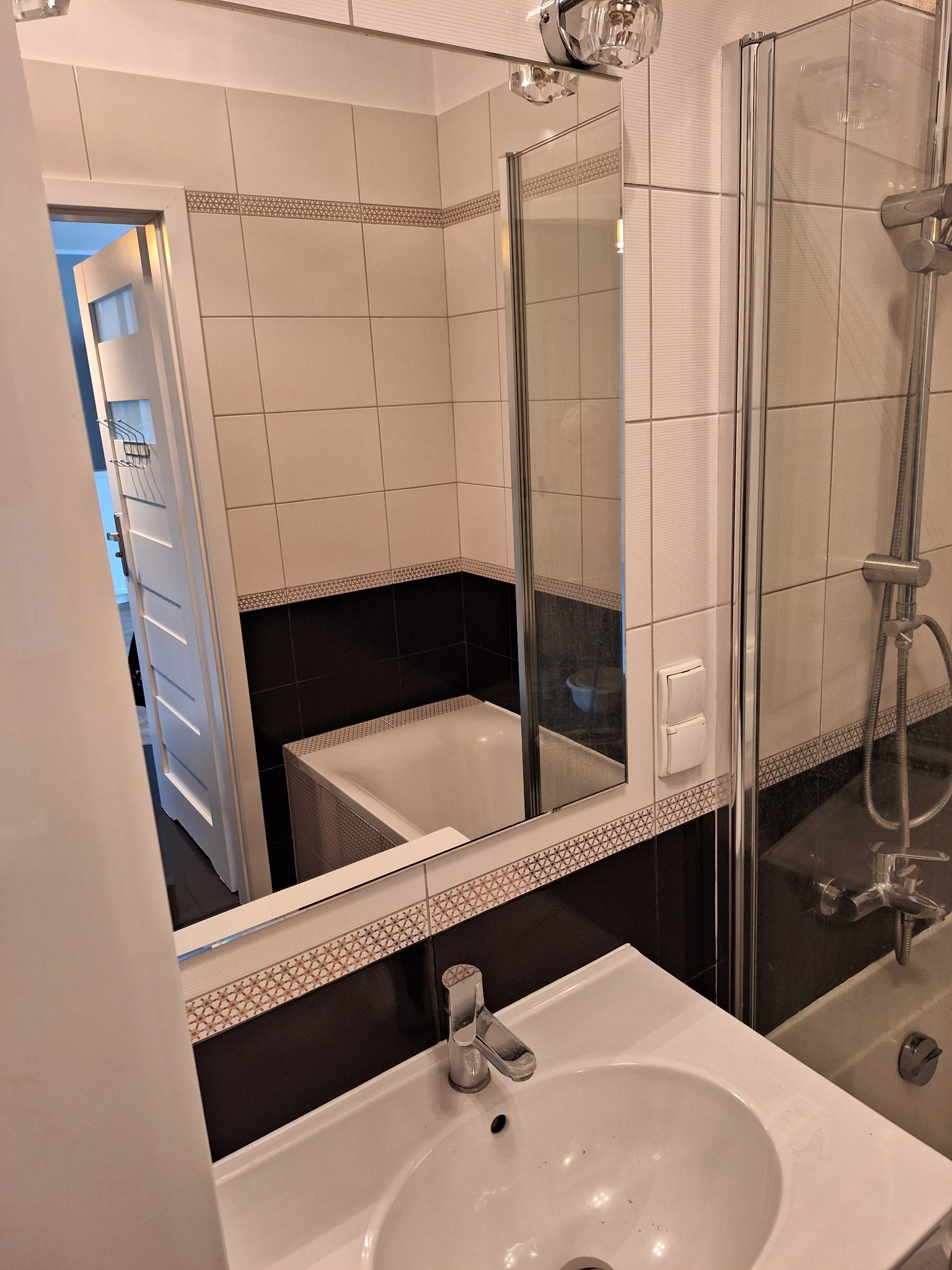 Umywalka z kranem w łazience z białymi i czarnymi płytkami, widok na kabinę prysznicową i lustro odbijające wannę.