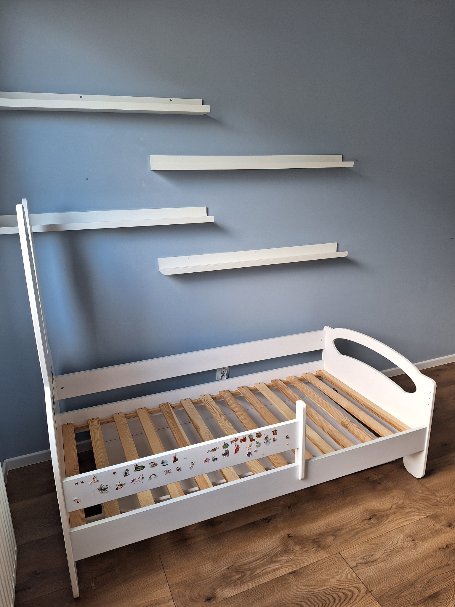 Białe łóżko dziecięce z naklejkami, drewnianym stelażem i trzema półkami na szarej ścianie, ustawione na parkiecie.
