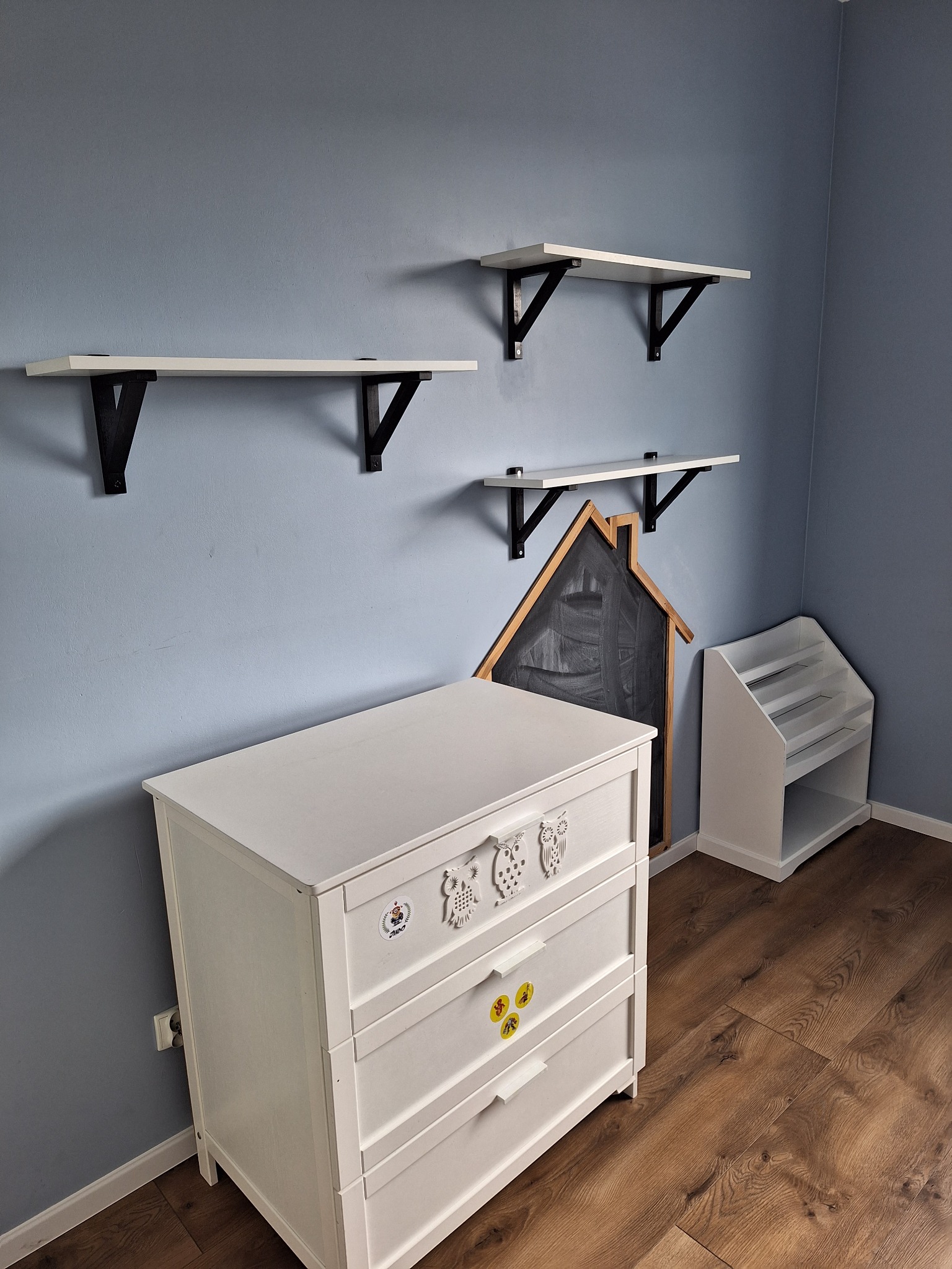 Pokój dziecięcy z białymi meblami: komoda z aplikacjami sów, regał na książki, tablica w kształcie domku oraz półki ścienne z czarnymi wspornikami na tle błękitnej ściany i drewnianej podłogi.