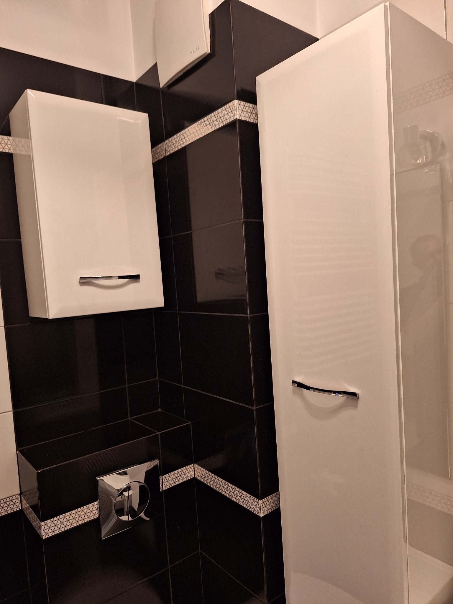 Ukośne ujęcie fragmentu łazienki z czarno-białymi płytkami, wiszącą białą szafką z chromowanym uchwytem, wysoką białą szafką przy prysznicu i chromowanym przyciskiem spłuczki.