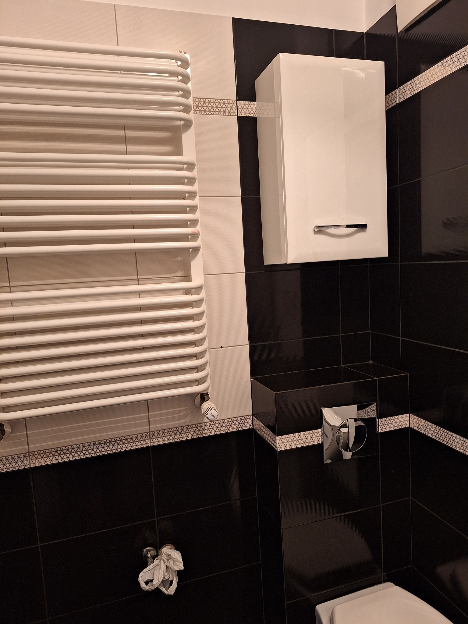 Biała łazienka z czarnymi akcentami: grzejnik drabinkowy, szafka nad toaletą i dekoracyjne płytki z geometrycznym wzorem.