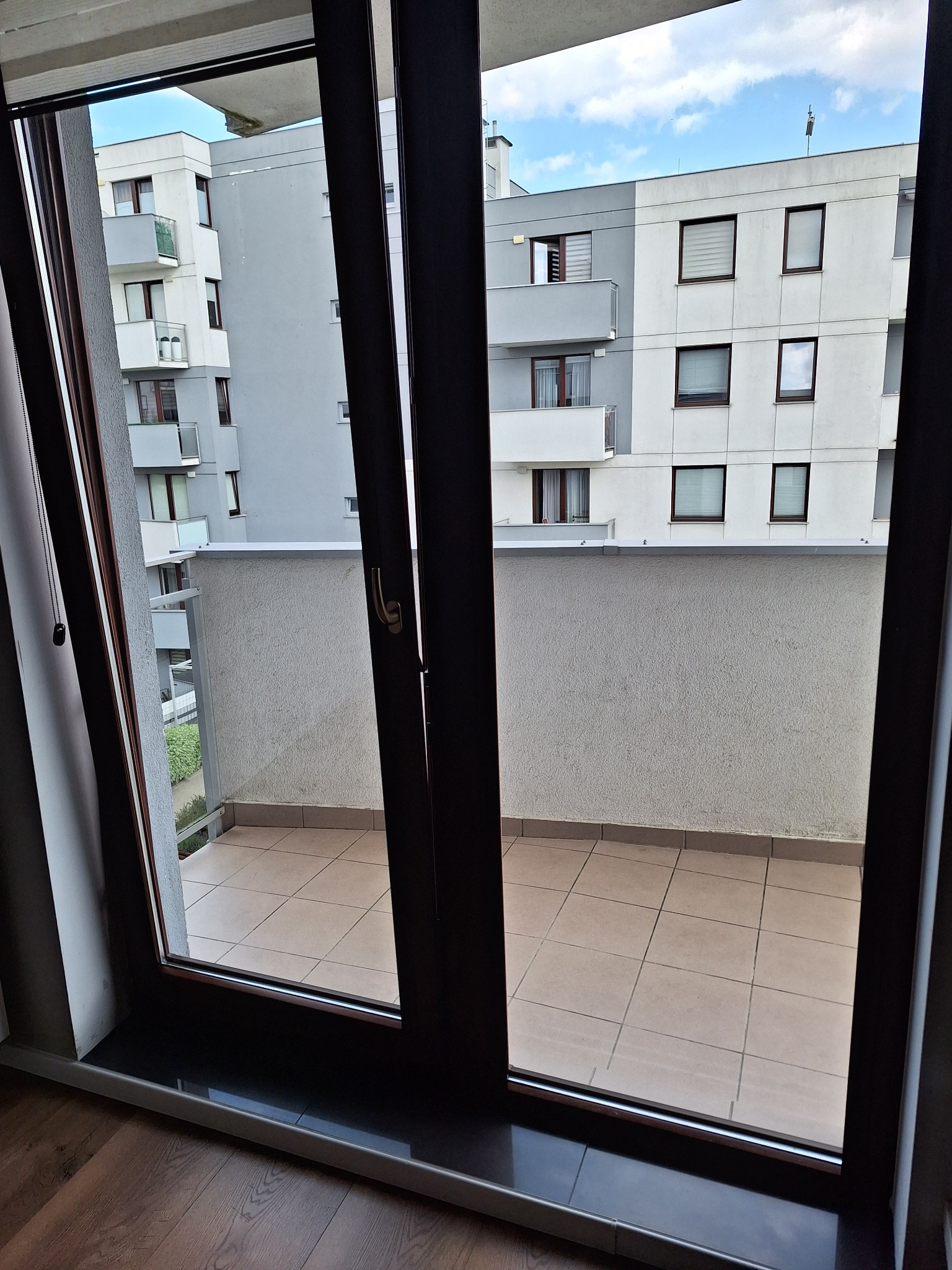 Widok z wnętrza mieszkania na balkon z beżowymi płytkami i szarym murem, w tle budynek wielorodzinny z balkonami i oknami, częściowo zasłoniętymi roletami.