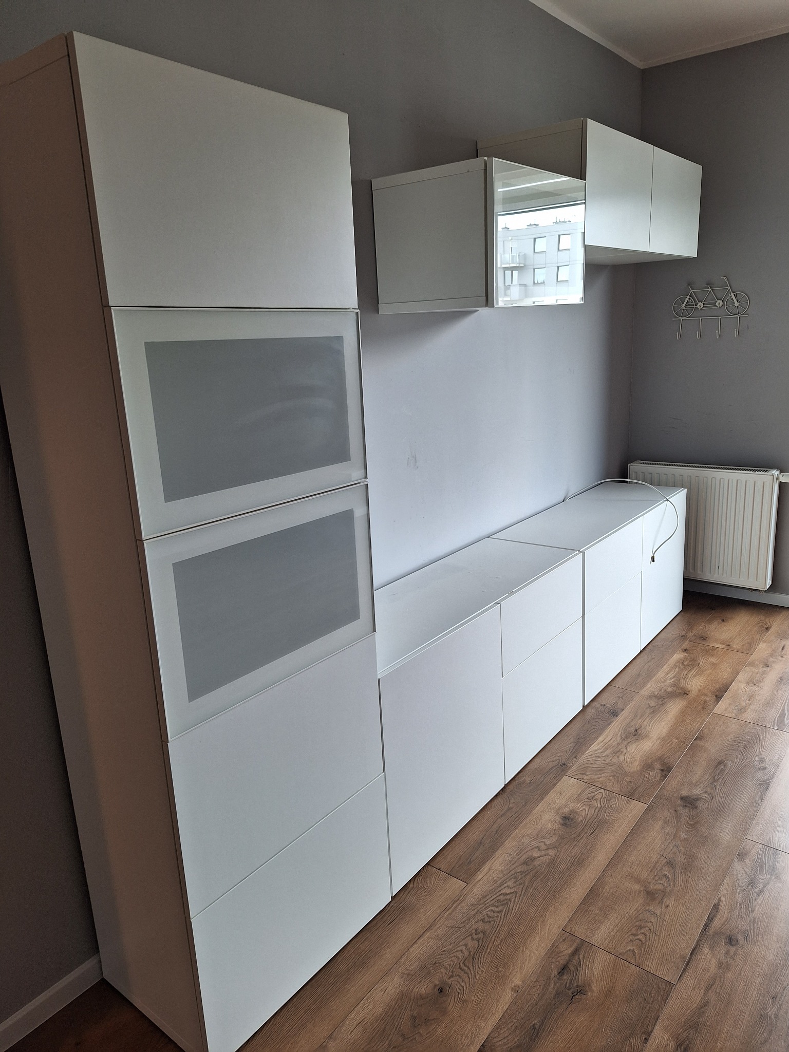 Jasne, nowoczesne meble modułowe z przeszklonymi frontami, drewniana podłoga, szara ściana z dekoracją rowerową i grzejnikiem w tle.