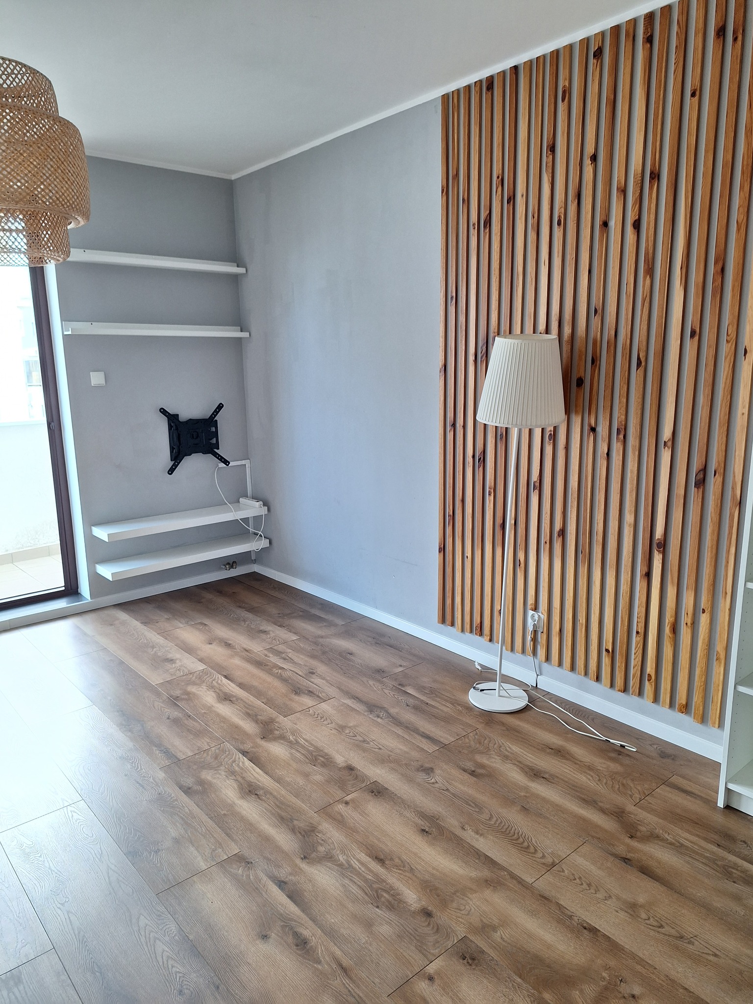 Pusty pokój po remoncie z drewnianą ścianką działową, podłogą laminowaną, zamontowanym uchwytem na telewizor i półkami na ścianie.