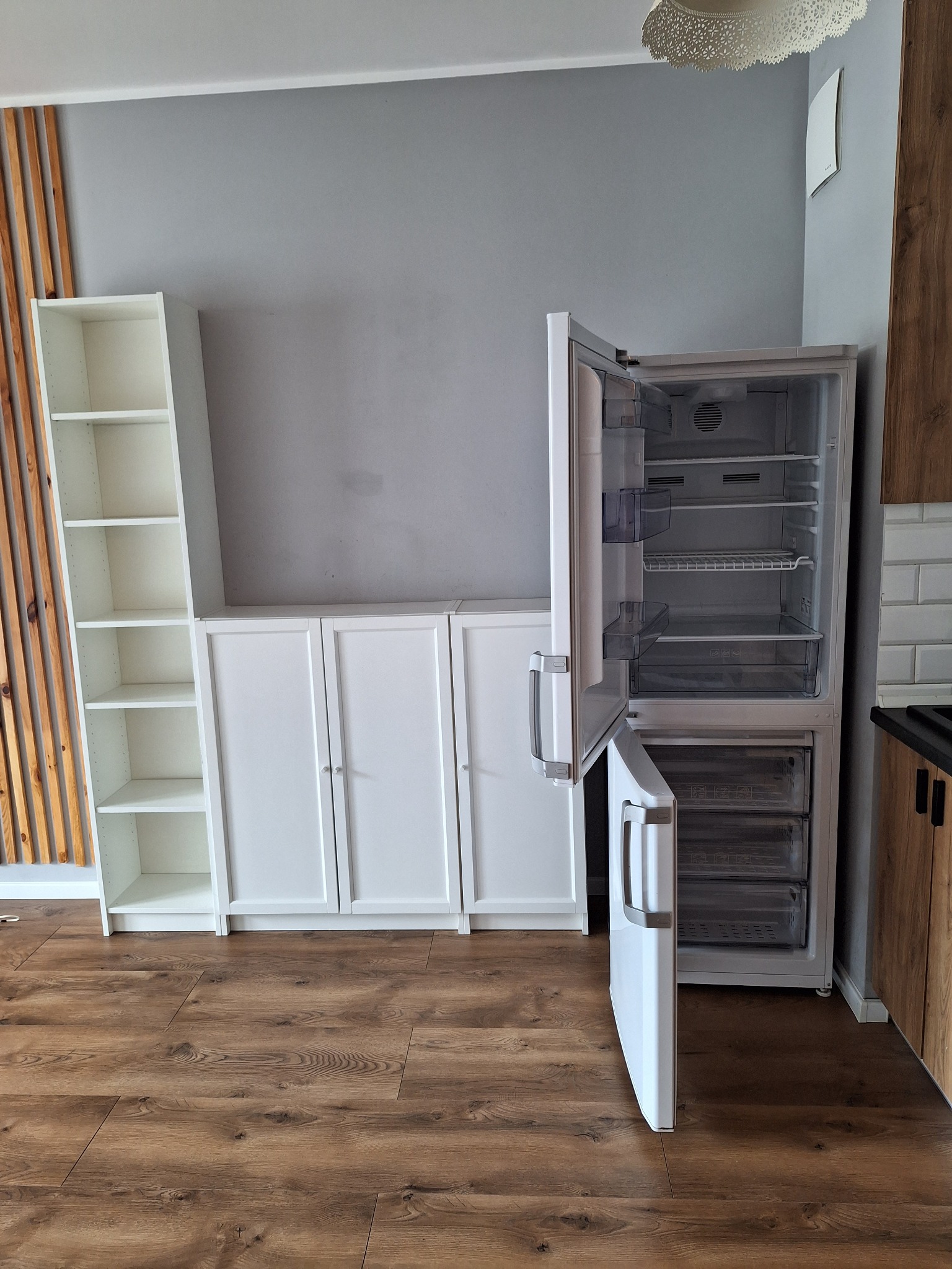 Częściowo opróżniona, biała lodówka z otwartymi drzwiami w mieszkaniu z drewnianą podłogą, białymi szafkami i półkami.