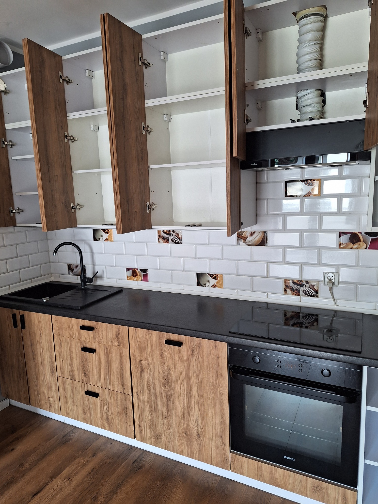 Nowoczesna kuchnia z drewnopodobnymi szafkami, czarnym blatem i białymi kafelkami z motywami kawowymi, szafki górne otwarte ukazujące wnętrze.