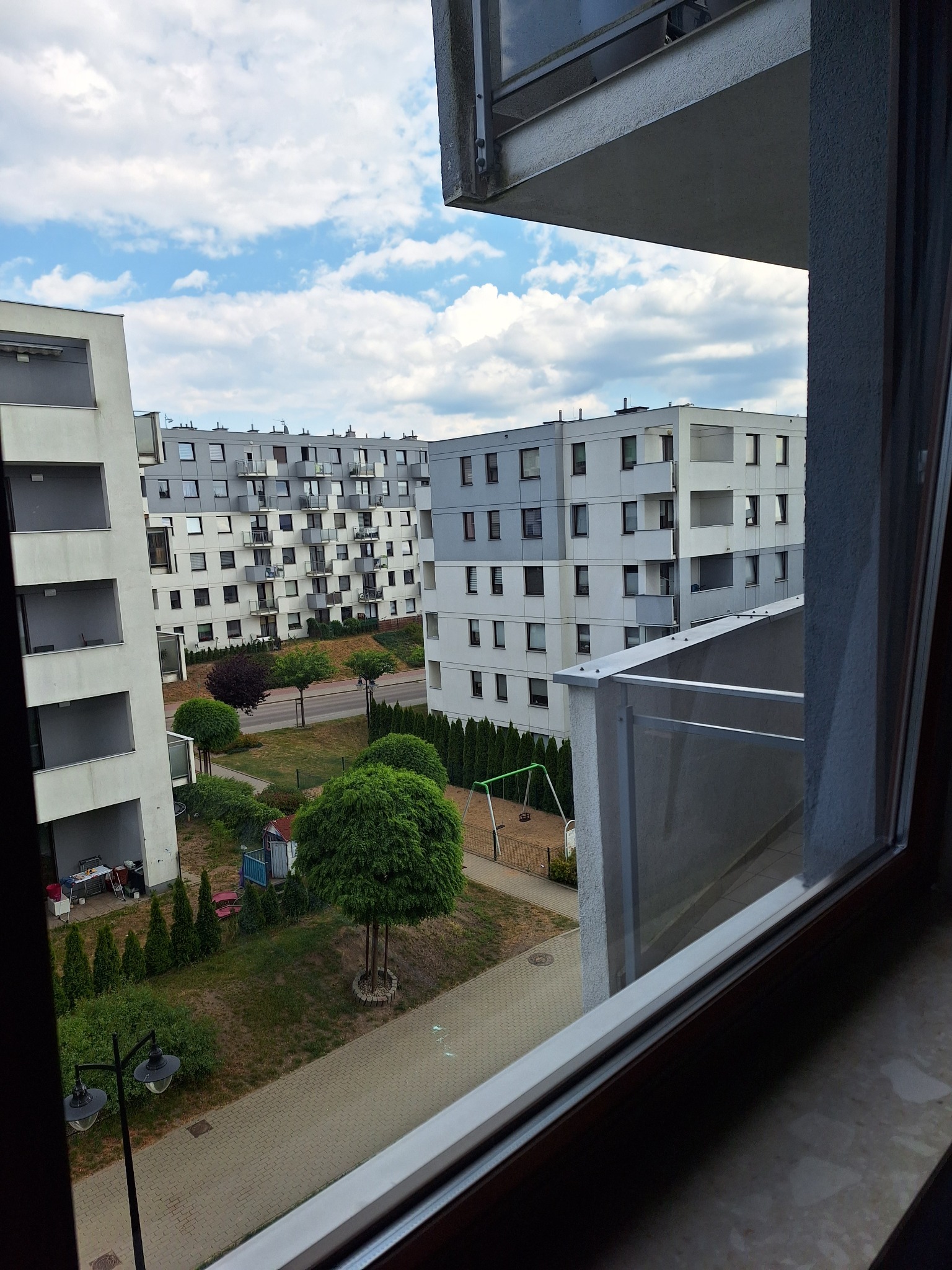 Widok z okna na osiedle bloków z placem zabaw i zielenią, częściowo przesłonięty ramą okienną.