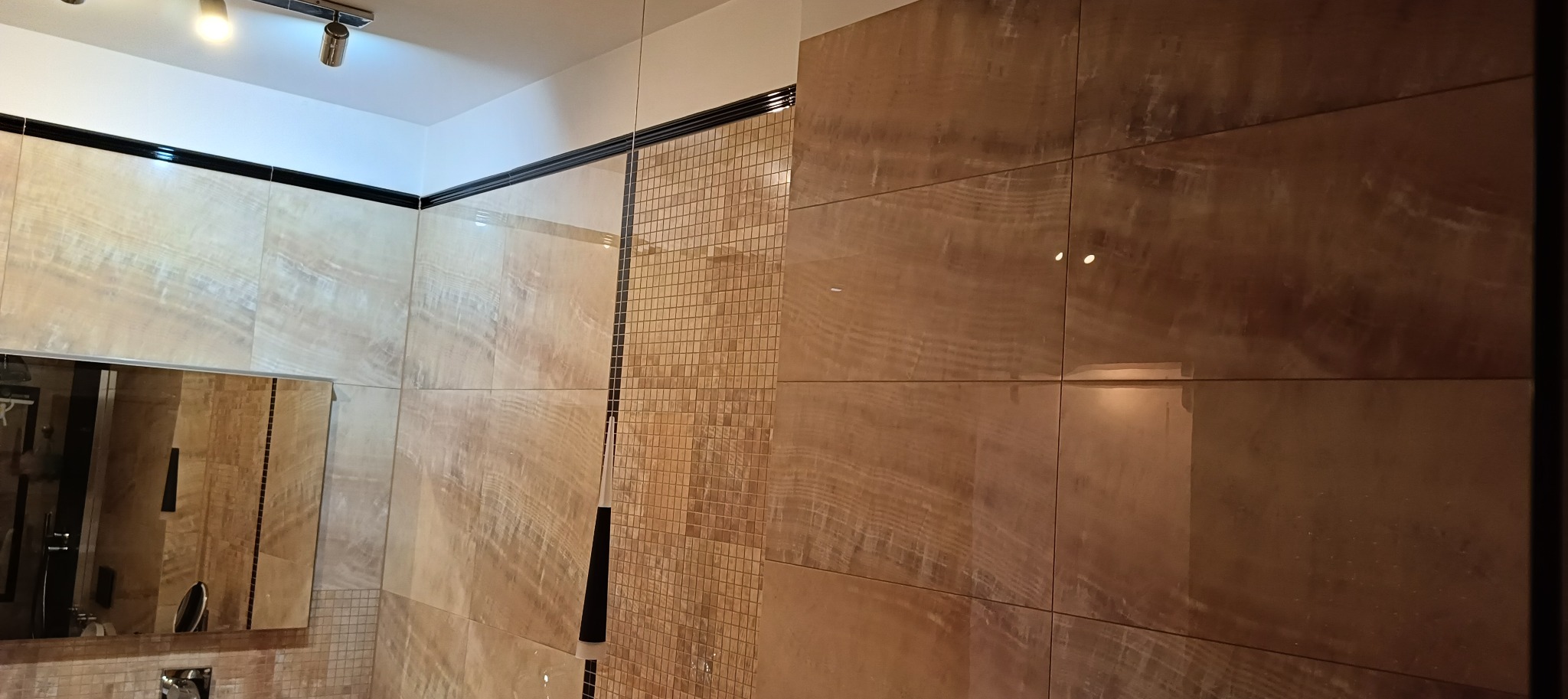 Wykończona łazienka z dużymi, beżowymi płytkami imitującymi marmur na ścianach, mozaiką w strefie prysznica i czarnymi akcentami wykończeniowymi.