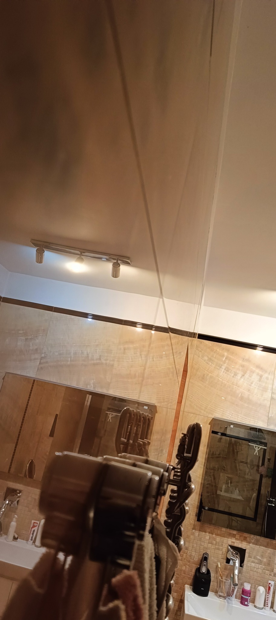 Ściany wykończone płytkami imitującymi drewno w łazience, widoczne oświetlenie sufitowe i odbicie w lustrze, z perspektywy osoby trzymającej wieszak na ręczniki.