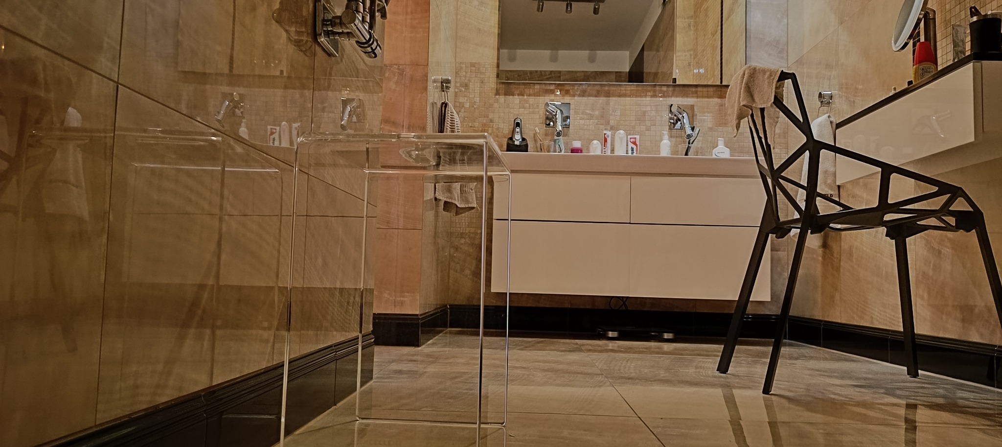 Nowoczesna łazienka z umywalką nablatową, lustrem i krzesłem o geometrycznej konstrukcji, wykończona płytkami w odcieniach beżu i brązu.