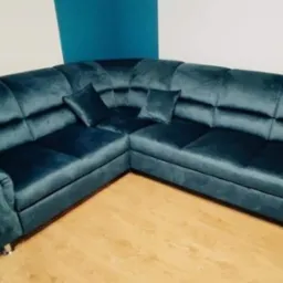 Narożnik tapicerowany w odcieniach szmaragdowej zieleni, z regulowanymi zagł&oacute;wkami i dwiema poduszkami, ustawiony na drewnianej podłodze, z fragmentem ściany w kolorze turkusowym w tle.