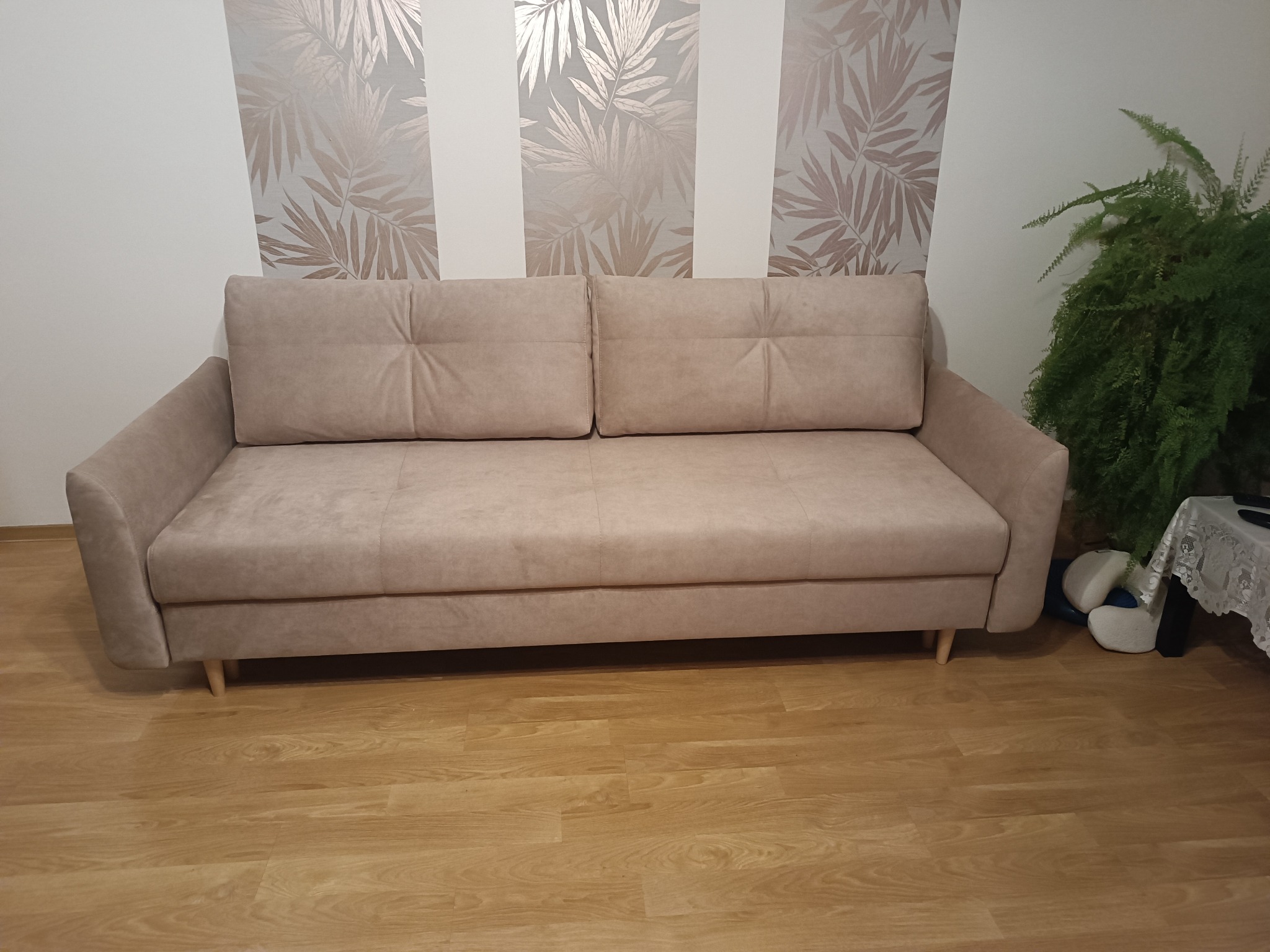 Beżowa sofa z dwoma poduszkami na oparciu, ustawiona na drewnianej podłodze, z wzorzystą tapetą w tle i zieloną rośliną po prawej stronie.