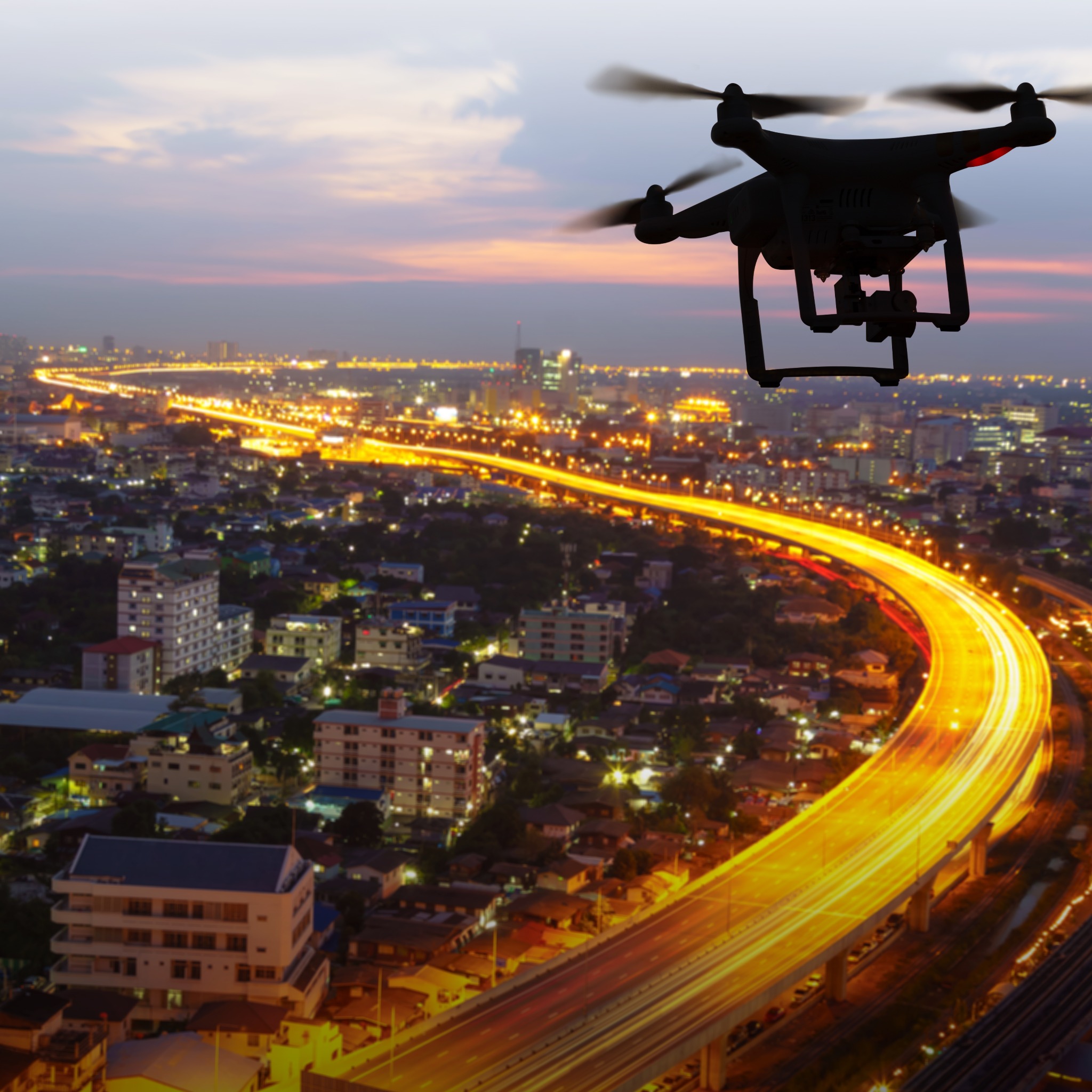 Sylwetka drona na tle nocnego miasta z długą ekspozycją świateł aut na wiadukcie.