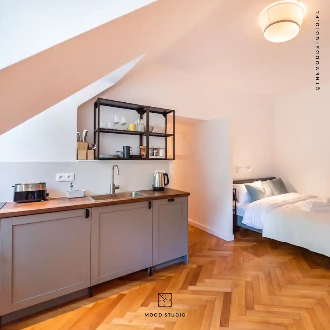 Aneks kuchenny z szarymi szafkami i drewnianym blatem w otwartej przestrzeni z sypialnią, podłoga w jodełkę, skośny sufit.