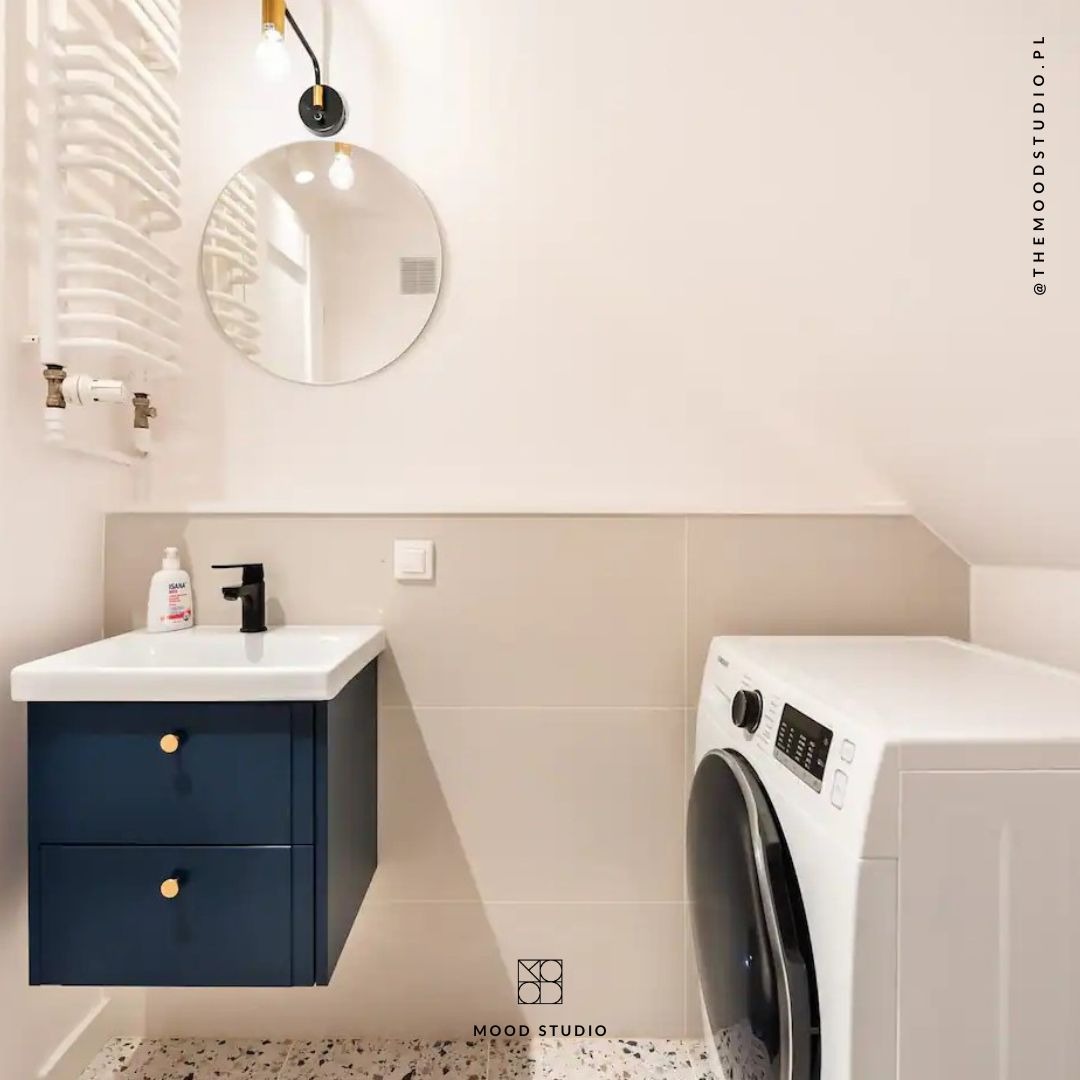 Nowoczesna łazienka z umywalką nablatową, szafką w kolorze granatowym z okrągłymi, złotymi uchwytami, okrągłym lustrem i białą pralką.