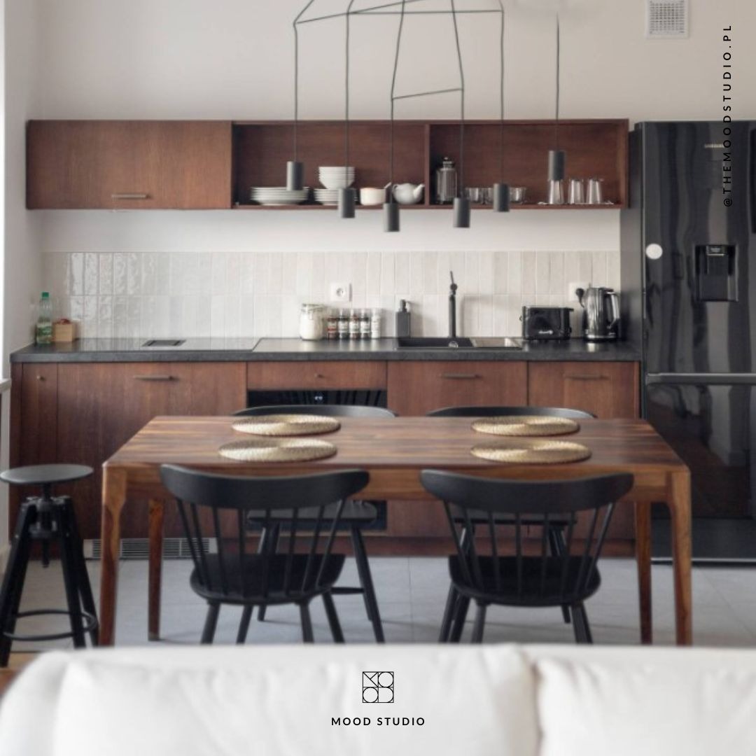 Kuchnia w stylu retro z drewnianymi szafkami, czarnym blatem i lodówką, stół z czarnymi krzesłami i okrągłymi podkładkami, dekoracyjna lampa wisząca.