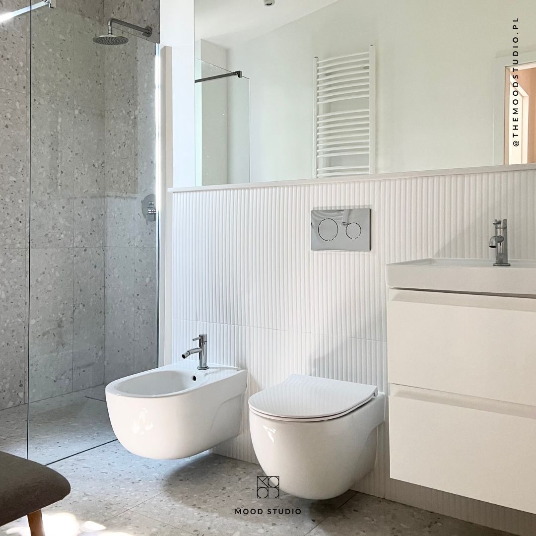 Jasna łazienka z kabiną prysznicową typu walk-in, wiszącą toaletą i bidetem, białą umywalką z szafką oraz grzejnikiem drabinkowym, wykończona płytkami lastryko na podłodze i szarymi płytkami...