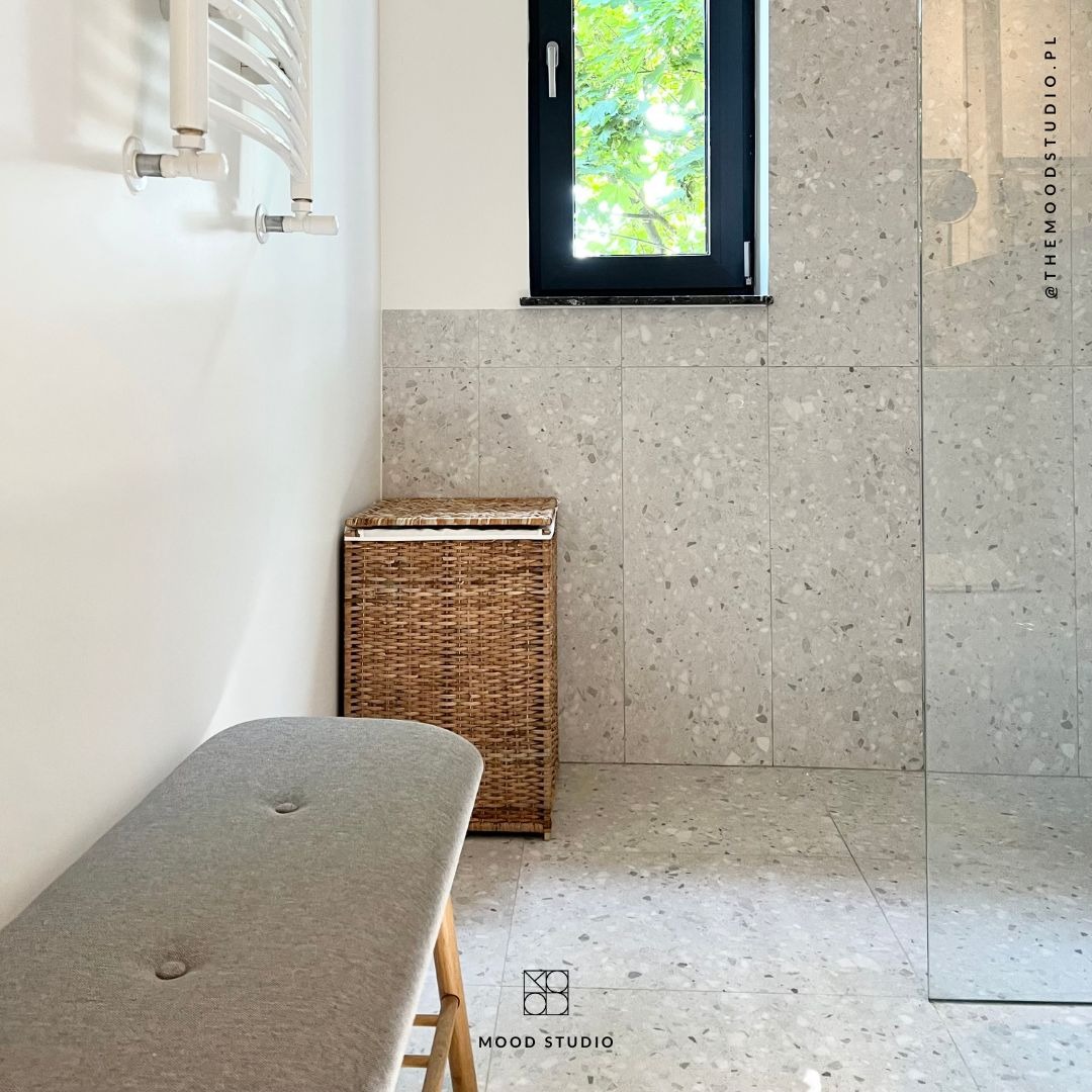 Jasna łazienka w stylu minimalistycznym z szarymi płytkami lastryko, wiklinowym koszem na pranie i szarym siedziskiem z dwoma guzikami.