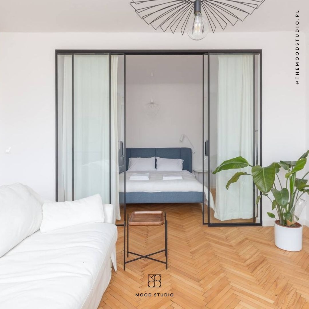 Salon z widokiem na sypialnię oddzieloną przesuwnymi, szklanymi drzwiami w czarnej ramie. Parkiet w jodełkę, biała sofa i roślina doniczkowa.