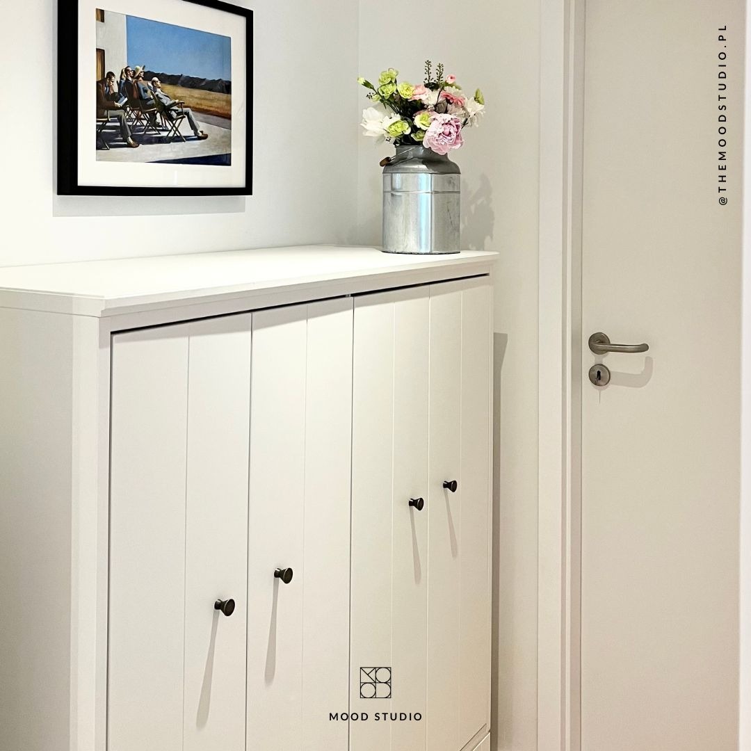 Biała szafka z czterema drzwiczkami i czarnymi gałkami, na której stoi metalowy dzbanek z bukietem kwiatów, nad szafką wisi obraz w czarnej ramie, obok widoczna biała ściana i drzwi.