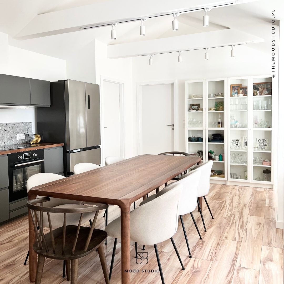 Jasne wnętrze jadalni z drewnianym stołem otoczonym krzesłami, szare szafki kuchenne z granitowym blatem, lodówka ze stali nierdzewnej, białe regały z przeszklonymi drzwiami, oświetlenie punktowe...