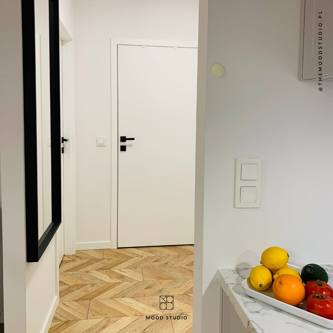 Jasne wnętrze przedpokoju z parkietem w jodełkę, białymi drzwiami z czarnymi klamkami oraz lustrem w czarnej ramie; na blacie widoczna misa z owocami i warzywami.