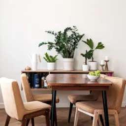 Jadalnia z drewnianym stołem, czterema krzesłami z beżową tapicerką, półką z roślinami doniczkowymi, karafką, świecznikami i misą z zielonymi winogronami na tle białej ściany i parkietu w jodełkę.