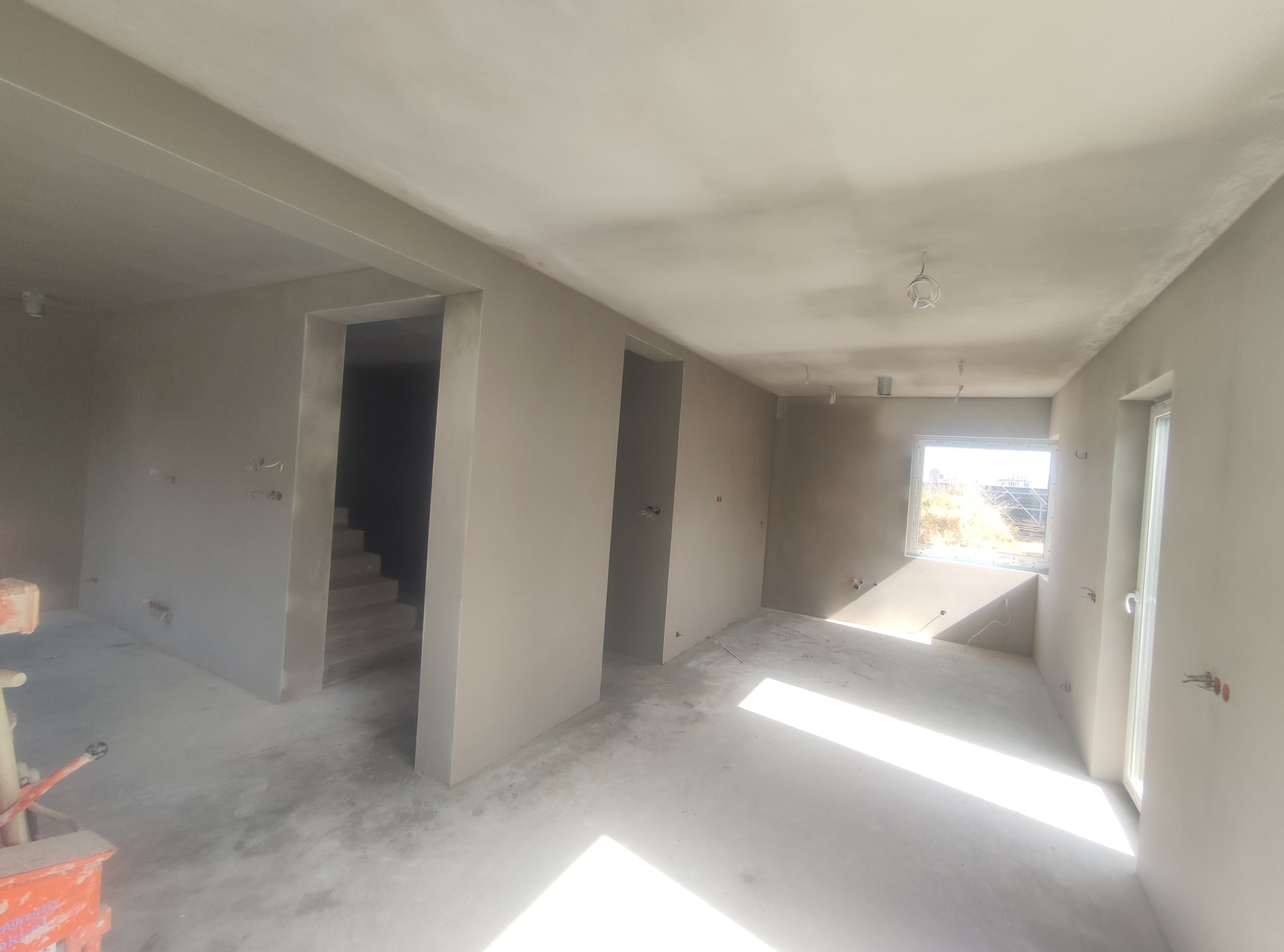 Wnętrze w stanie deweloperskim po nałożeniu szarej warstwy wykończeniowej na ściany i sufity, widoczne przygotowane otwory okienne i drzwiowe oraz schody w głębi.