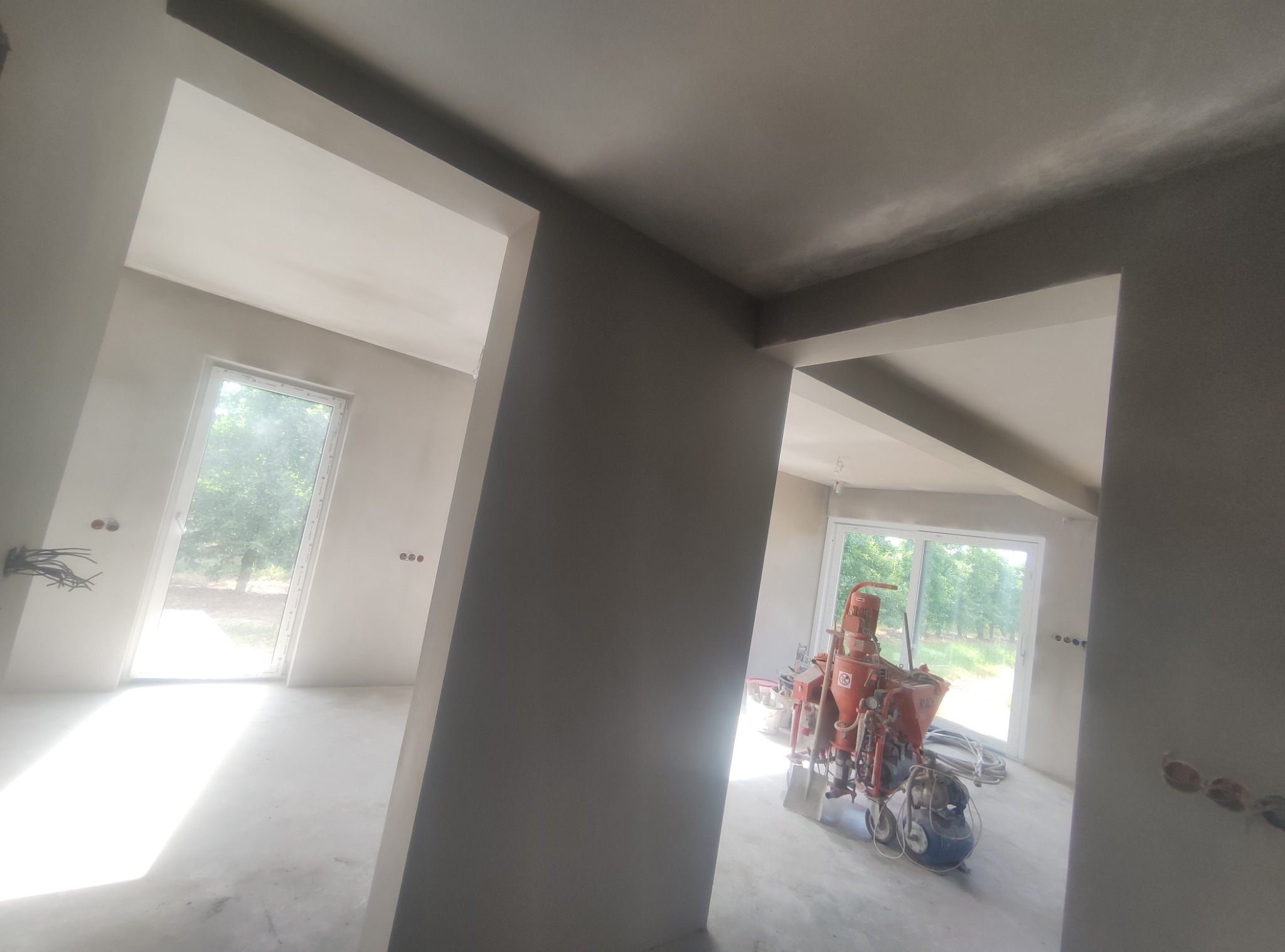 Wnętrze w stanie deweloperskim z maszyną do tynkowania, świeżo położone tynki na ścianach i suficie, przygotowane pod dalsze prace wykończeniowe, widoczne okna z zielenią na zewnątrz.