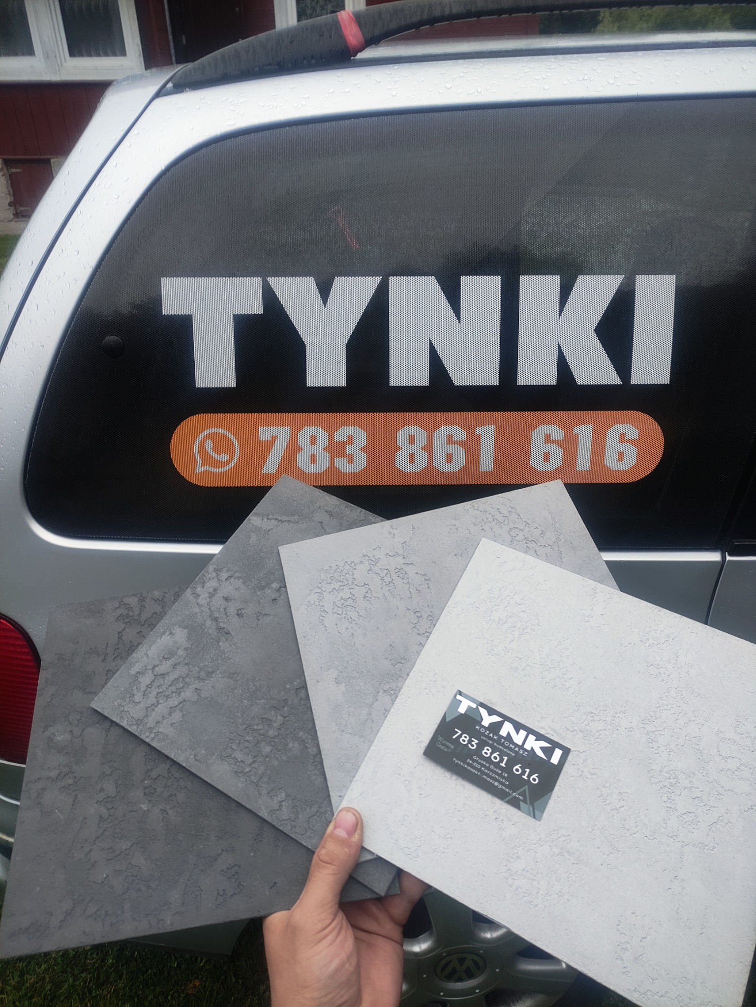 Próbki faktur w odcieniach szarości prezentowane na tle samochodu z naklejką reklamową firmy budowlanej.