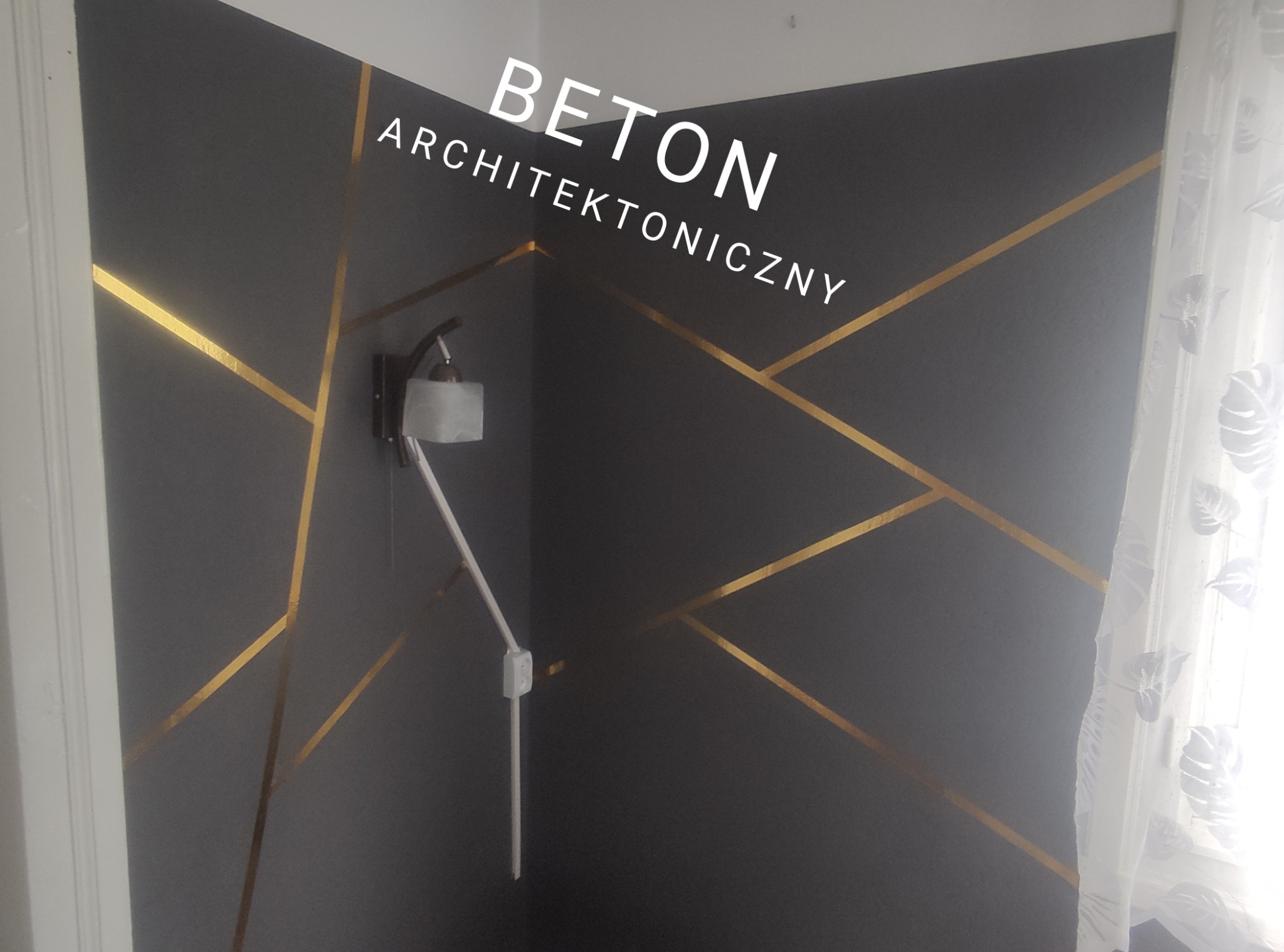 Ściana w ciemnoszarym odcieniu z efektem betonu architektonicznego, zdobiona złotymi, geometrycznymi liniami, oświetlona kinkietem z białym abażurem. W tle widoczna biała, wzorzysta firanka.