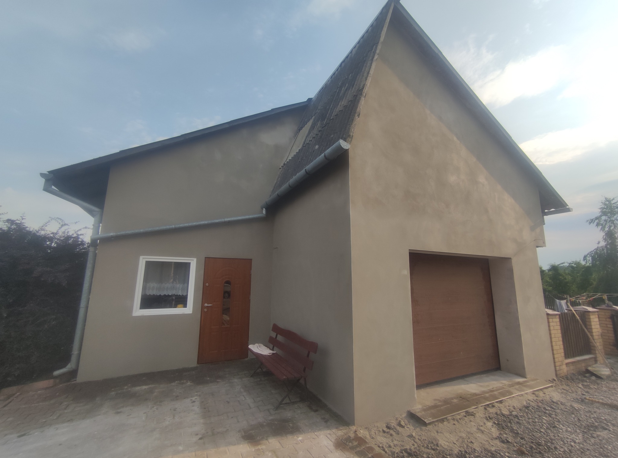 Świeżo otynkowana elewacja budynku z brązowymi drzwiami garażowymi i wejściowymi, z widocznym fragmentem dachu z eternitu i ławką przed wejściem.