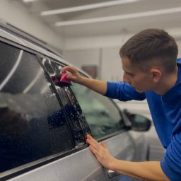 FREEDOM DETAILING MYJNIA - Pracownik w niebieskiej bluzie aplikuje folię na szybę srebrnego auta, używając różowego aplikatora. Widoczne krople wody.