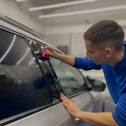 Pracownik w niebieskiej bluzie aplikuje folię na szybę srebrnego auta, używając różowego aplikatora. Widoczne krople wody.