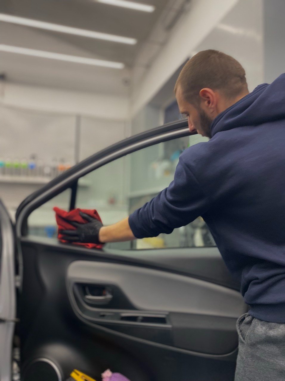 Mężczyzna w czarnych rękawiczkach czyści szybę auta czerwoną ściereczką w jasnym wnętrzu myjni. Widoczne detale wnętrza samochodu i oświetlenie sufitowe.