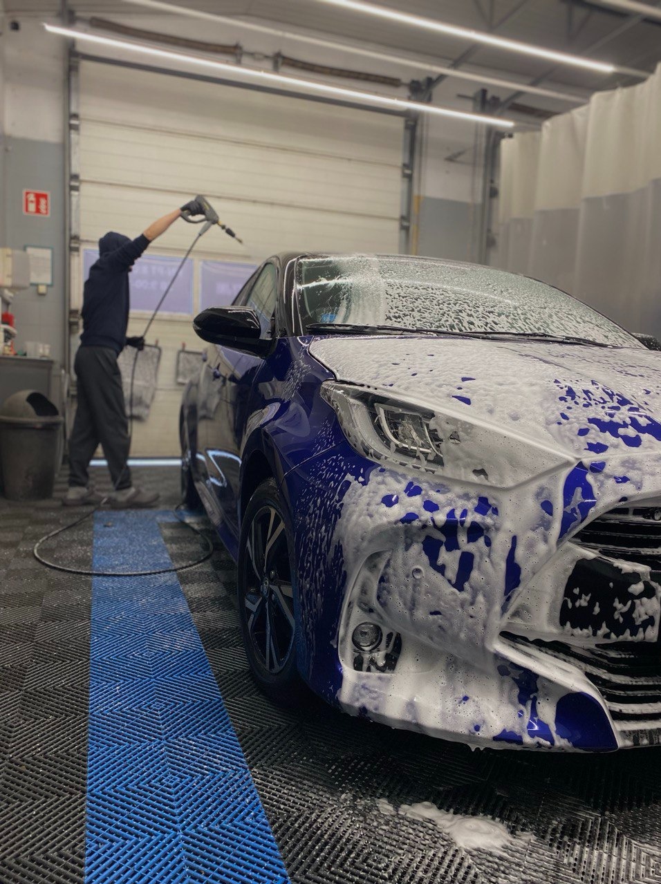 Mycie niebieskiego auta Yaris w myjni ciśnieniowej. Widoczna piana na karoserii, pracownik z lancą i charakterystyczna niebieska linia na podłodze.