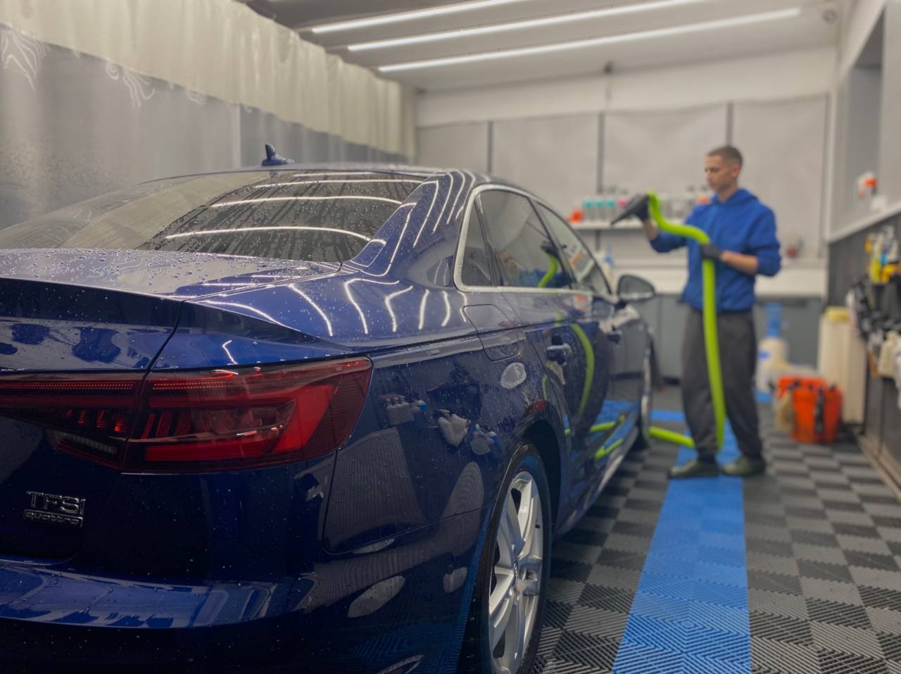 Błyszczący, mokry, granatowy samochód Audi po myciu w myjni, w tle pracownik suszy auto za pomocą węża z dyszą. Widoczne krople wody na karoserii.