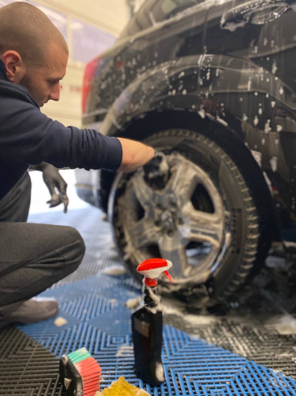 Czyszczenie felgi czarnego auta z użyciem piany i szczotki. Widoczny pracownik w rękawicach, butelka z rozpylaczem i gąbka na podłodze.